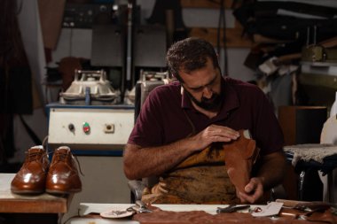 Becerikli bir zanaatkar dağınık bir tezgahta oturur, kahverengi deriyi titizlikle şekillendirir. Loş atölye deri, aletler ve yakınlarda bir çift bitmiş ayakkabıyla dolu..