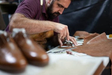Odaklanmış bir zanaatkar, yakındaki bitmiş ayakkabıları olan bir tezgahta deri parçaları buduyor, el sanatlarını yakalıyor ve hareketli bir stüdyoda geleneksel ayakkabı yapma sanatını icra ediyor..