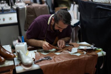 Yetenekli bir zanaatkar, ölçüm aletleri, makas ve yapıştırıcıyla çevrili kahverengi deri üzerinde dikkatle çalışır. Sahne, canlı bir atölyede titiz bir el işi ve geleneksel ayakkabı yapımını ön plana çıkarıyor..