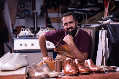 Yetenekli bir kunduracı, deri bir önlük giymiş, deri ayakkabı ve aletlerle çevrili olarak dağınık bankında oturur. Gülümsüyor, el işçiliği ve küçük işletme sanatının gururunu yansıtıyor..