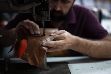 Odaklanmış bir zanaatkar, kahverengi deriyi dikiş makinesinden geçirir, hassas dikişler atar. El yapımı deri işçiliği, kalitesi ve bakım için el yapımı aletler..
