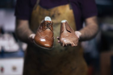 Lekeli önlüklü yetenekli bir ayakkabıcı bitmemiş bir ayakkabının yanında parlak kahverengi bir ayakkabı tutar. Geleneksel ayakkabı yapımını ve el yapımı ayakkabı sanatını yakalar..