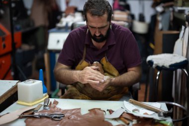 Yetenekli bir deri işçisi meşgul bir masada oturur ve elleriyle deri bir parça şekillendirir. Bitmiş ve dayanıklı bir yaratılışa konsantre olurken araçlar ve kırıntılar yakınlarda uzanıyor..