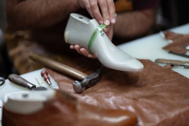 Yetenekli bir ayakkabıcı, yeşil bir mezura kullanarak protez ayakkabı boyutunu kontrol eder. Çalışma tablosundaki deri parçalar ve aletler, dikkatli montaj ve onarım teknelerini vurguluyor.