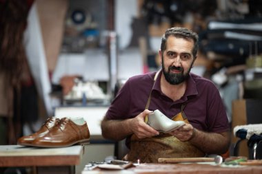Önlük giyen yetenekli bir zanaatkar yoğun bir atölyede beyaz deri bir ayakkabı kullanıyor. Ilık ışık odaklanmasını, banktaki aletleri ve bitirmeye hazır kahverengi bir deri ayakkabıyı vurgular..
