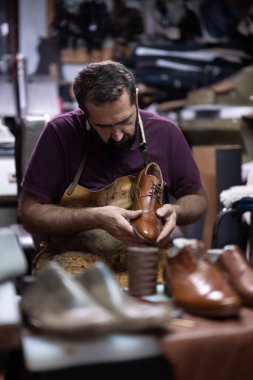 Lekeli önlük giymiş deneyimli bir ayakkabıcı, yoğun bir atölyede aletler ve deriyle çevrili kahverengi deri ayakkabıyı inceler ve arka planda tamamlanmış ve beklenen çiftleri görür..