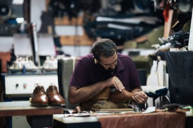 Yetenekli bir ayakkabı tamircisi, el yapımı ayakkabılara ve ayakkabı tamirine adanmışlığını gösteren alet ve malzemelerle dolu bir atölyede kahverengi bir deri botu dikkatlice cilalar..