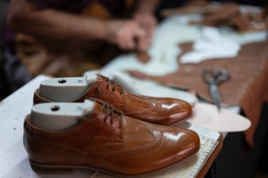 Bir çift cilalı kahverengi deri ayakkabı, meşgul bir ayakkabı tamirhanesinde dayanır. Alet ve malzemeler arka planda bulanıklaşıyor, onarım, işçilik ve geleneksel erkek giyim tarzları.