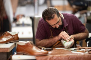 Yetenekli bir zanaatkar yoğun bir atölyede kahverengi deri ayakkabıları şekillendirmeye ve bitirmeye konsantre olur. Aletler, deri kırıntıları ve bitmiş çiftler çalışma alanını çevreleyerek, geleneksel