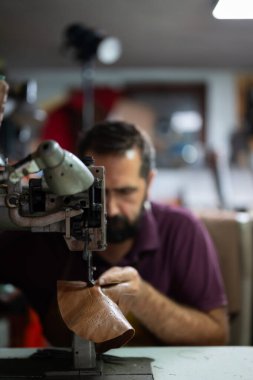 Sakallı bir adam, yoğun bir atölyede, işçilik, deri işçiliği ve geleneksel ayakkabı yapımı gibi ağır bir dikiş makinesine kahverengi bir deri botu elle dikerken konsantre olur..