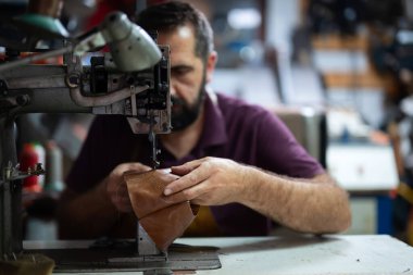 Odaklanmış bir deri ustası deri bir panele ağır endüstriyel dikiş makinesinde rehberlik ediyor. Atölye atmosferi pratik ve zanaatkar hissettiriyor. Yetenekli işçiliği vurguluyor.