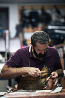 Mor gömlek ve önlük giyen yetenekli bir zanaatkar bankta çekiç kullanarak deri bir çizmeyi dikkatlice yamalar ve şekillendirir. Dağınık atölye çalışması geleneksel ayakkabı tamir becerilerini gösteriyor..