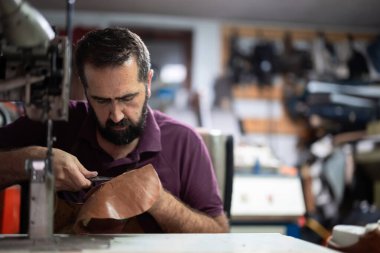 Yoğun bir atölyede çalışan odaklı bir ayakkabı tamiratı için el dikişleri atan bir ayakkabı tamircisi. Etrafını saran aletler, deri kırıntıları ve makineler, işçilik, sabır ve geleneksel