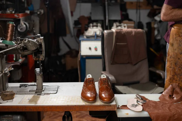Dikiş sehpasında cilalanmış kahverengi Oxford ayakkabılarının bulunduğu, iplik, deri ve el işçiliği ve marifetli el yapımı işçiliği taşıyan alet dolu bir çevreyle çevrili bir ayakkabı atölyesi sahnesi..