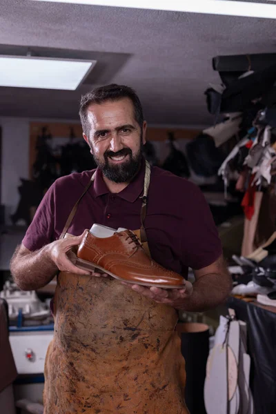 Deri önlüklü yetenekli bir zanaatkar, deri, alet ve malzemelerle dolu bir atölyede parlak kahverengi bir deri ayakkabı bulundurur. İşçiliği, kalitesi ve gururu ön plana çıkarır.