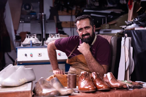 Yetenekli bir kunduracı, deri bir önlük giymiş, deri ayakkabı ve aletlerle çevrili olarak dağınık bankında oturur. Gülümsüyor, el işçiliği ve küçük işletme sanatının gururunu yansıtıyor..