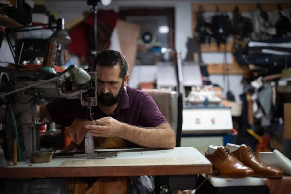 Deri dikiş makinesinde çalışan bir adam dağınık bir atölyede, kahverengi deri parçaları şekillendirirken yakındaki ayakkabıların cilalanmasını sağlıyor. Sahne, zanaat, odaklanma ve küçük ölçekli ayakkabı yapımı içeriyor..