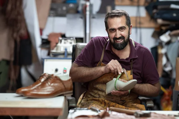 Neşeli bir ayakkabıcı, meşgul bir deri atölyesinde oturur ve bir ayakkabıyı bant ile ölçer. Önlük giyiyor ve el yapımı deri ayakkabılar üzerinde çalışıyor. Etrafı aletlerle çevrili ve kahverengi bir ayakkabı..