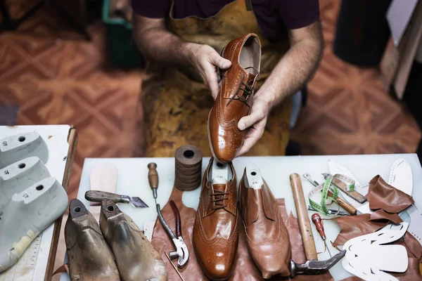 Yetenekli bir zanaatkar cilalanmış kahverengi deri ayakkabılar, el aletleri, iplik makaraları ve kırıntılarla çevrili. Elinde bir ayakkabı tutarak, geleneksel ayakkabı imalatına odaklanmış bir atölyede gösteri yapıyor..