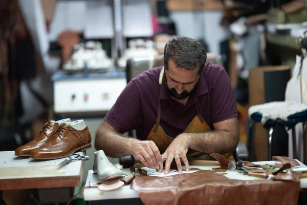 Önlük giyen yetenekli bir zanaatkar kahverengi ayakkabılar için deri parçalar üzerinde titizlikle çalışır. Aletler ve artıklar masaya yayılmış, odak, beceri ve geleneksel ayakkabı imalatı