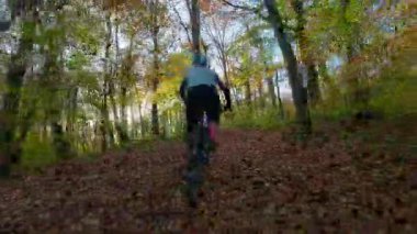 Tek başına bir dağ bisikletçisi, güzel bir sonbahar gününde yapraklarla kaplı orman yolunda manzaralı bir gezinti yapar. Altın güneş ışığı, doğanın neşesini vurgulayarak, canlı sonbahar yapraklarını aydınlatır.