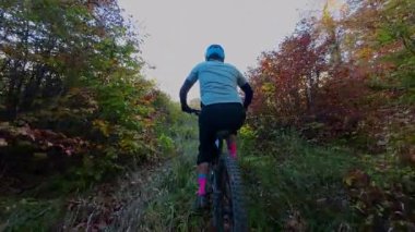Tek başına bir dağ bisikletçisi, güzel bir sonbahar gününde yapraklarla kaplı orman yolunda manzaralı bir gezinti yapar. Altın güneş ışığı, doğanın neşesini vurgulayarak, canlı sonbahar yapraklarını aydınlatır.