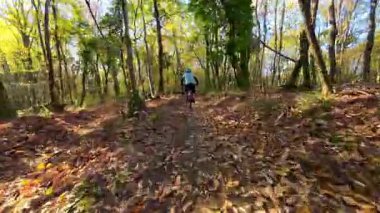 Aktif bir insan, sonbaharda manzaralı bir orman patikasında eğlenceli bir dağ bisikleti yolculuğundan hoşlanır. Güneş ışığı renkli gölgeliklerden süzülür, düşen yapraklarla kaplı yolu aydınlatır.