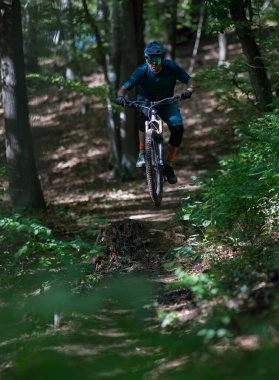 Bir kask ve koruyucu giysi giyen odaklanmış bir sürücü, toprak orman yolunda küçük bir atlayışı temizler, atletizm ve bereketli ağaçlar ve benekli ışık ile açık hava macerası sergiler..