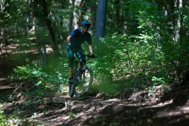 Yalnız bir dağ bisikletçisi tozlu bir orman patikasında miğfer ve atletizm kıyafetleri giyerek geziniyor. Yeşil yeşilliklerle çevrelenmiş olan sahne, doğada odaklanma, hız ve açık hava macerası taşır..
