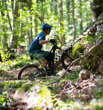 Kask takan odaklanmış bir dağ bisikletçisi, güneşli bir orman yolunda tam kapasite süspansiyon bisikleti sürüyor, açık hava sporu, spor, spor ve macera sergiliyor yeşil ağaçlar ve kayalık arazi arasında..