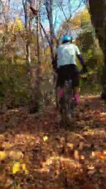 Mavi dişli bir sürücünün dikey FPV görüntüsü elektrikli bir dağ bisikleti üzerinde ağaçlık bir patikada, miğferinde güneşli, yapraklı bir orman sahnesinde benekli ışık ve yosunlu zemin ile geziniyor..