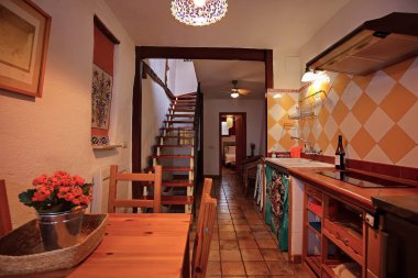 Sıcak, davetkar bir iç mekan, içinde ahşap bir yemek masası, saksı bitkisi, üst kata çıkan merdivenler, turuncu elmas kaplama ve yumuşak ortam aydınlatması olan parlak bir mutfak..