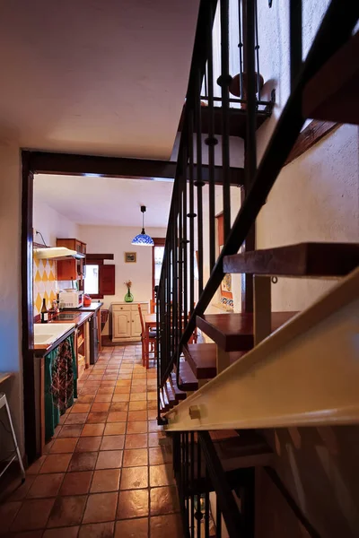 Kiremit kaplı bir zemin, solda bir mutfak, metal parmaklıklı ahşap bir merdiven ve sıcak ışıkla yıkanmış bir arka yemek alanı..