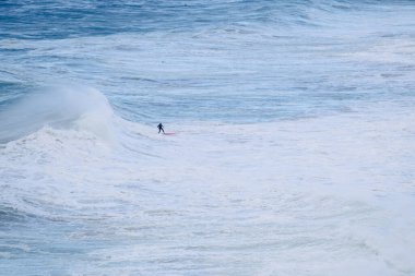 Yalnız bir sörfçü, engin mavi su ve beyaz sörf ile kıvrımlı bir okyanus dalgasında dengesini korur. Dinamik, maceralı sahne. Yetenek, odaklanma ve doğanın ham gücü..