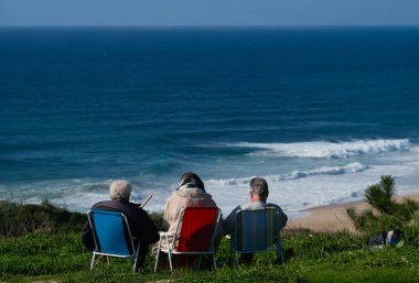 Üç son sınıf öğrencisi kumlu bir plajın yukarısındaki çimenli bir tepede, açık bir gökyüzünün altında dalgaların gelişini seyrediyor. Huzurlu bir birliktelik ve deniz kenarında bir eğlence anı..
