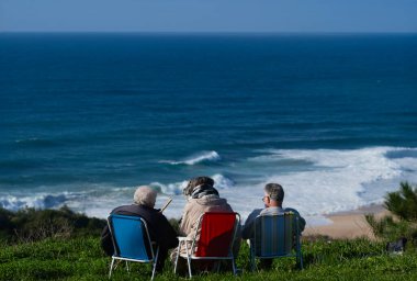 Üç yaşlı arkadaş çimenli bir tepede renkli bahçe sandalyelerinde rahatlar, deniz kıyısında sakin ve manzaralı bir an için mavi dalgaların gelişini izlerler..