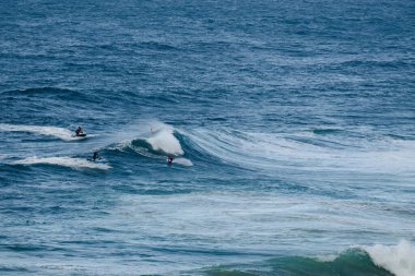 Sörfçüler beyaz köpükle güçlü mavi dalgalara binerler, açık okyanusta hareket, denge ve heyecan yakalarlar.