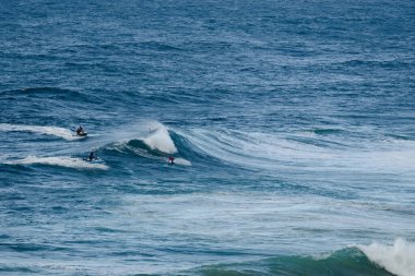 Bir grup sörfçü, arkalarında beyaz köpük düşerken sörf tahtalarında dengeli bir şekilde engin mavi okyanus boyunca güçlü dalgalara biniyor. Dinamik, enerjik spor tasvirleri macera temaları için mükemmel.