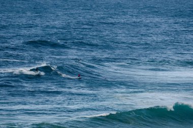 Dalgalar üzerinde süzülen sörfçüler uçsuz bucaksız mavi bir okyanusta süzülür. Enerji, hareket ve açık hava sporlarını gösterir. Biniciler beyaz köpüklerin arasında dengede durur..