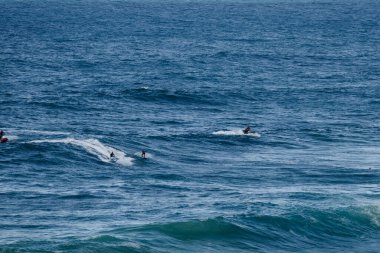 Bir grup sörfçü, açık okyanustaki enerjik mavi dalgalarda gezinerek paralel dalgalarda dolaşırken sprey ve beyaz köpük oluştururlar. Dinamik spor hareketi ve güneşli bir günde çekilmiş kıyı eğlencesi.