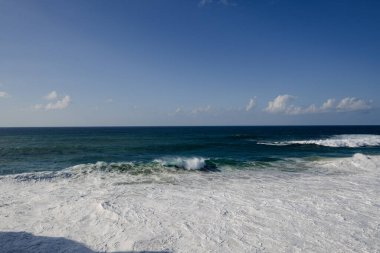 Derin mavi okyanusun sakin kıyı manzarası, dalgalar ve parlak bir gökyüzü. Geniş ufku olan doğa, seyahat veya rahatlama temaları için mükemmel.