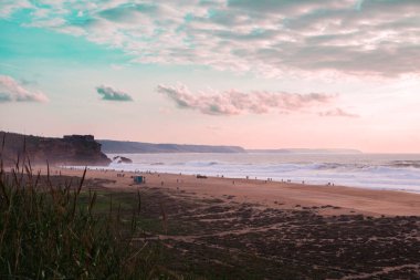 Sakin bir sahil manzarası, kumlu bir sahil, yumuşak dalgalar, pastel gökyüzü ve uzak uçurumlar. Ön plandaki çimenler manzarayı çerçevelerken, küçük insan grupları kıyıdan keyif alıyorlar..