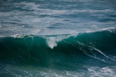 Açık denizde büyük bir turkuaz dalganın dramatik bir çekimi, beyaz köpük takip ediyor, canlı mavi-yeşil tonlar, okyanusun hareketini ve enerjisini yakalıyor..