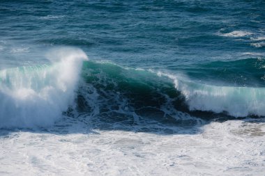 Turkuaz ibikli ve beyaz köpüklü, seyahat, doğa ve spor temalarına uygun enerji ve kıyı atmosferini taşıyan dramatik bir kıvrımlı okyanus dalgası sahile çarpar..