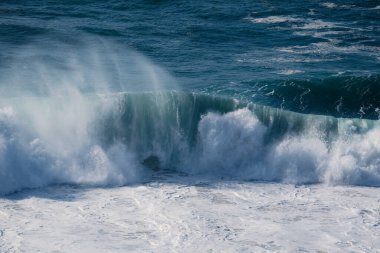 Yükselen bir okyanus dalgası kıvrılır ve çarpar, havaya sprey ve köpük gönderir, denizin enerjisini ve hareketini derin mavi tonlarda yakalar..
