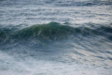 Dinamik deniz manzarası, kıyıya yakın köpüklü beyaz köpükle derin mavi bir okyanusta büyük dalgalar ve kıvrımlı dalgalar gösteriyor. Seyahat, doğa veya spor temaları için uygun enerjik doğal kıyı sahnesi.