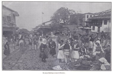 Geleneksel elbiseli insanların, meyve ve mal satan satıcıların ve tarihi bir Arnavut kasabasında hareketli bir kalabalığın bulunduğu kaldırımlı bir sokakta hareketli bir pazar sahnesi..