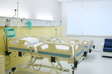 Bir hastane odasında, birkaç boş hastane yatağı, tıbbi ekipmanların yanı sıra küçük bir oturma alanı düzenlenir. Doğal ışık boşluğu doldurur ve sakin bir atmosfer yaratır..