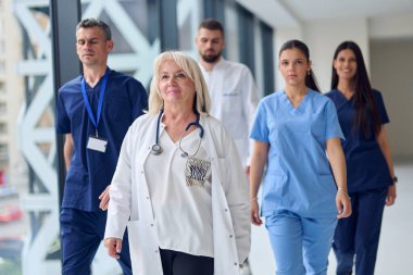 Çeşitli sağlık çalışanlarından oluşan bir grup hastane koridorunda takım çalışması ve profesyonellik sergileyerek birlikte yürüyor. İlerlerken üniforma giyerler ve odaklanmış ifadeler sergilerler..