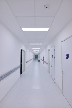 Hastanedeki temiz beyaz koridor çeşitli odaların kapılarını gösteriyor. Parlak floresan ışıkları mekanı aydınlatarak ziyaretçiler ve çalışanlar için sakin bir ortam yaratıyor..